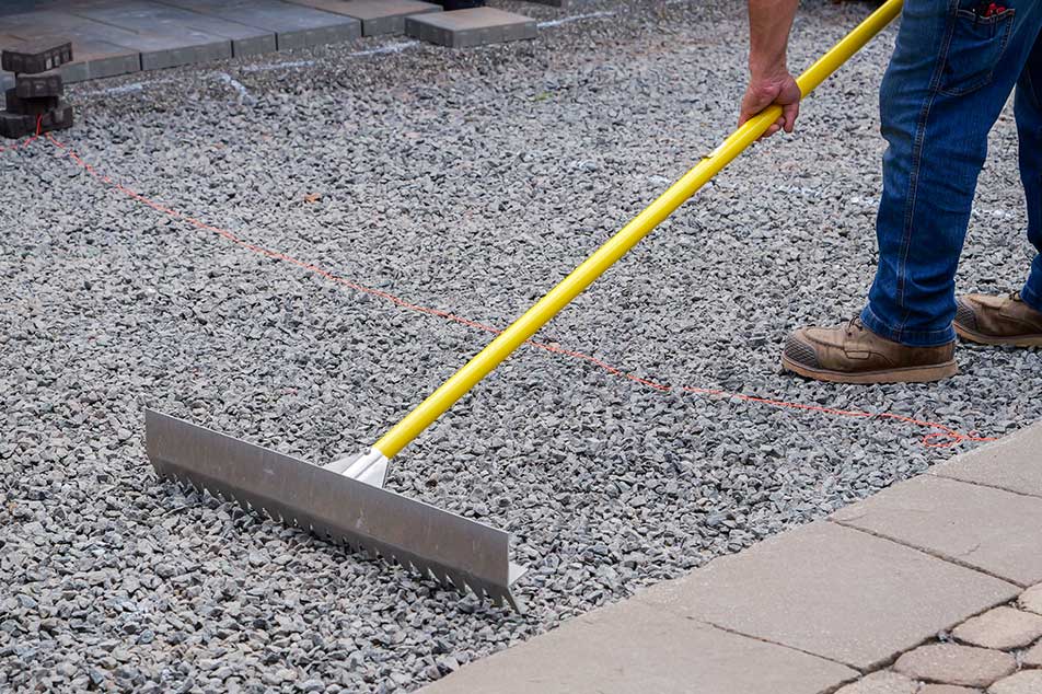 Person using a yellow tool on a gravel surface with a partially completed stone pathway.